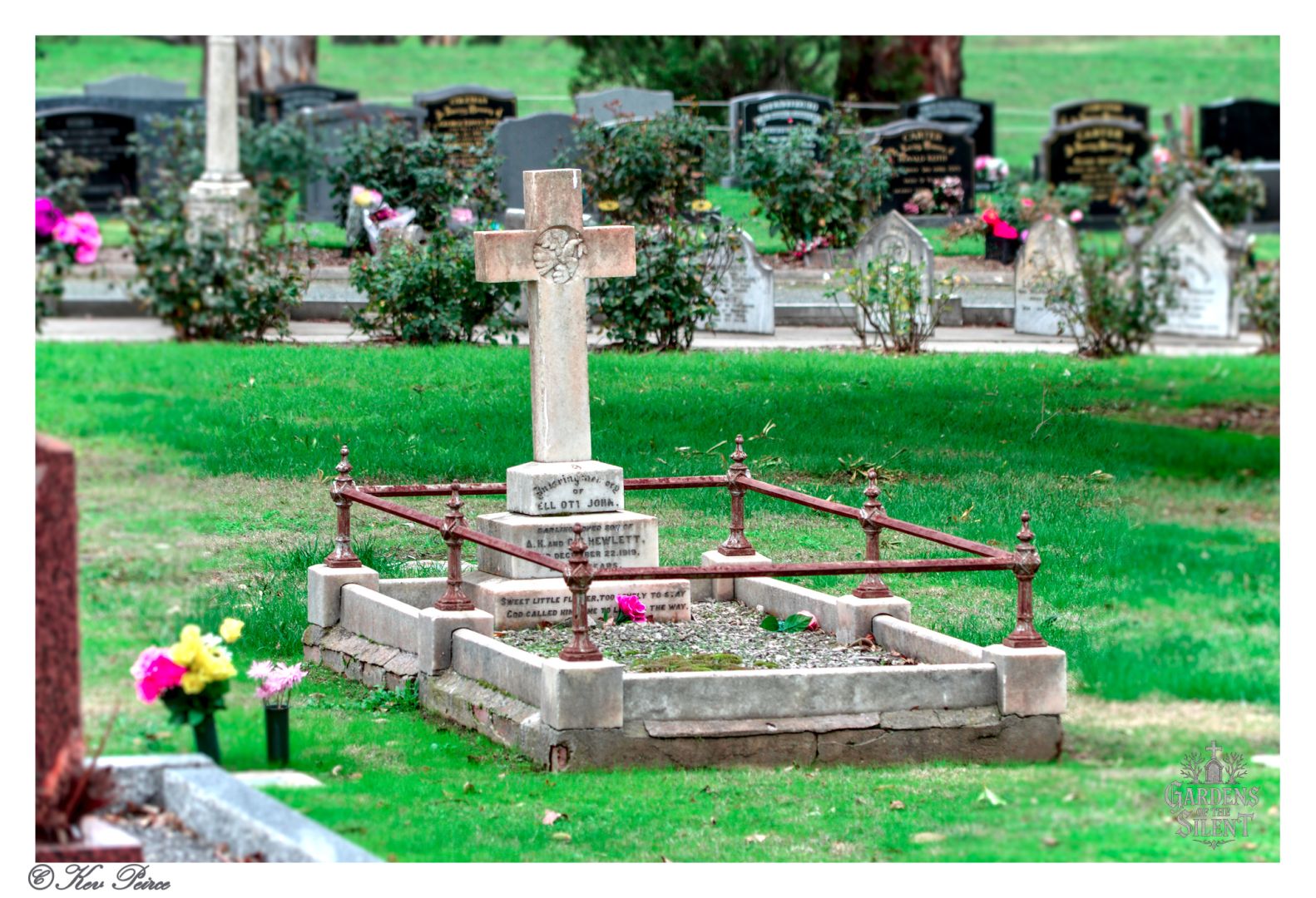 A headstone with a cross and a small metal fence around the plot at Gumeracha Cemetery.  The grave is in the foreground with other headstones and a grassy field behind it.
