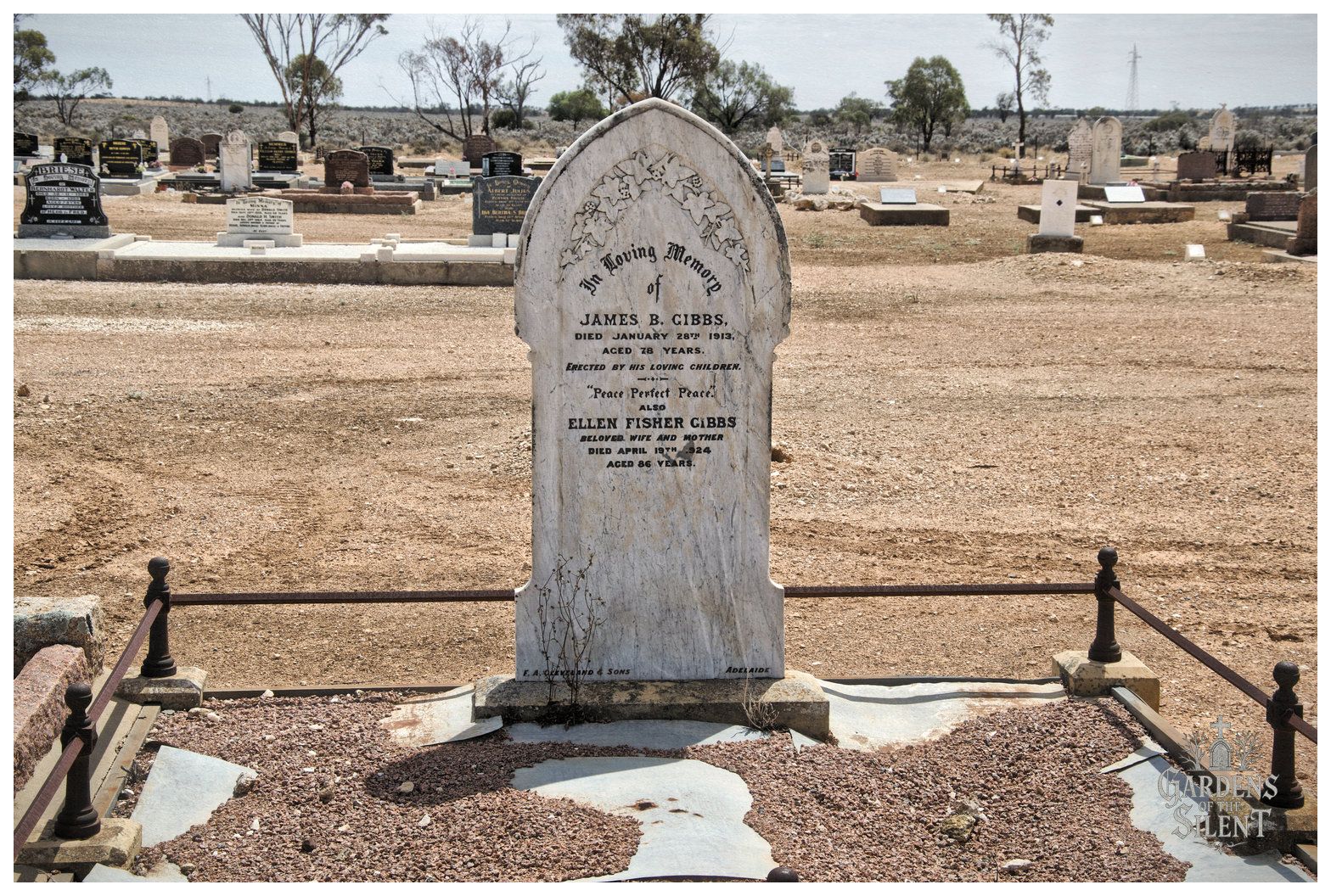 A colour landscape photograph featuring a tall, pointed white marble headstone decorated with carved ivy motifs, dedicated to James B. Gibbs and Ellen Fisher Gibbs. The headstone stands behind a low, dark iron fence enclosing a bed of reddish-brown gravel with cracked concrete. In the background, the sunlit, dusty landscape of the cemetery extends towards distant gum trees under an overcast sky.