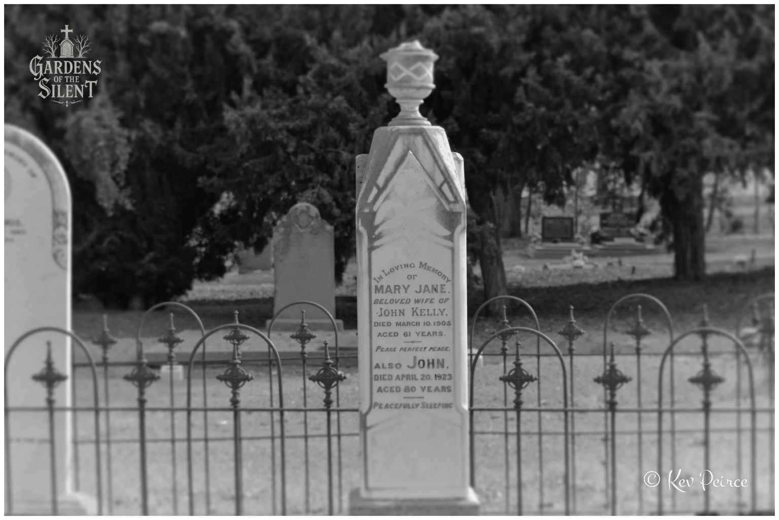 A black and white photograph capturing a tall marble obelisk topped with a decorative stone urn, dedicated to Mary Jane and John Kelly. The monument stands directly behind a low, scalloped iron plot fence. The surrounding cemetery grounds and background trees are gently blurred with a soft-focus depth of field.