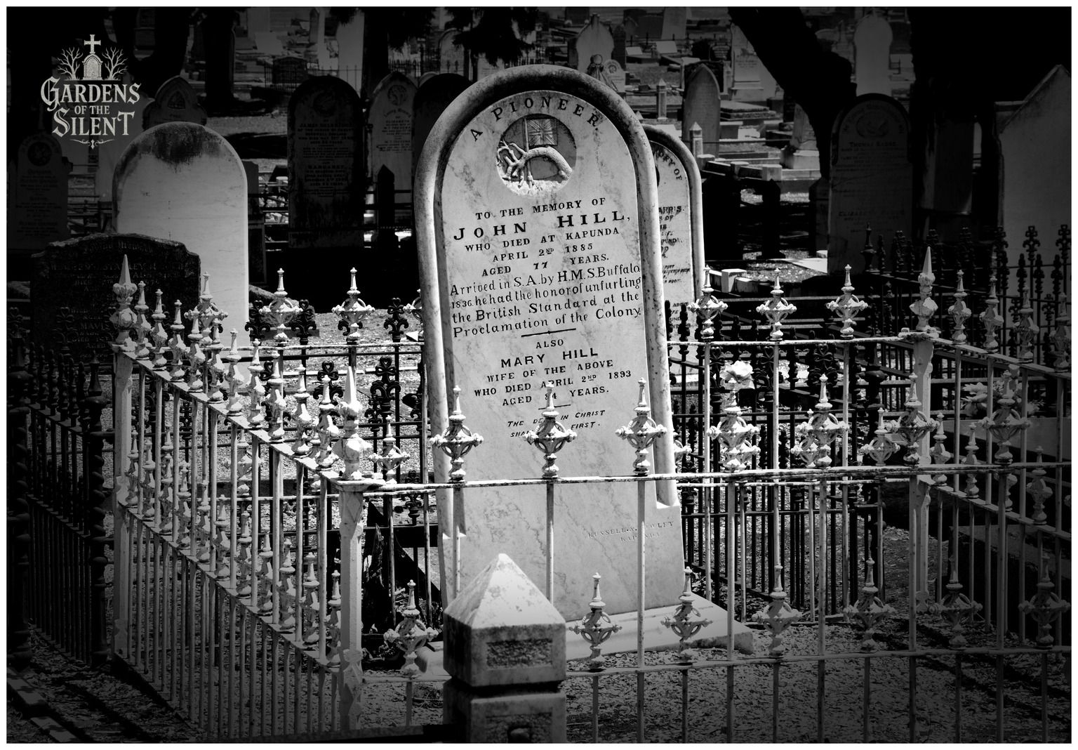 A black and white photograph focusing on an arched marble headstone dedicated to John Hill, a pioneer who arrived in South Australia in 1836 on the H.M.S. Buffalo. The top of the stone features a carved relief of a hand holding a flag. The grave is completely enclosed by an intricate, light coloured iron picket fence with decorative finials. Shadows stretch across the background where other headstones stand in the dark. A watermark reading 'Gardens of the Silent' is in the top left corner.