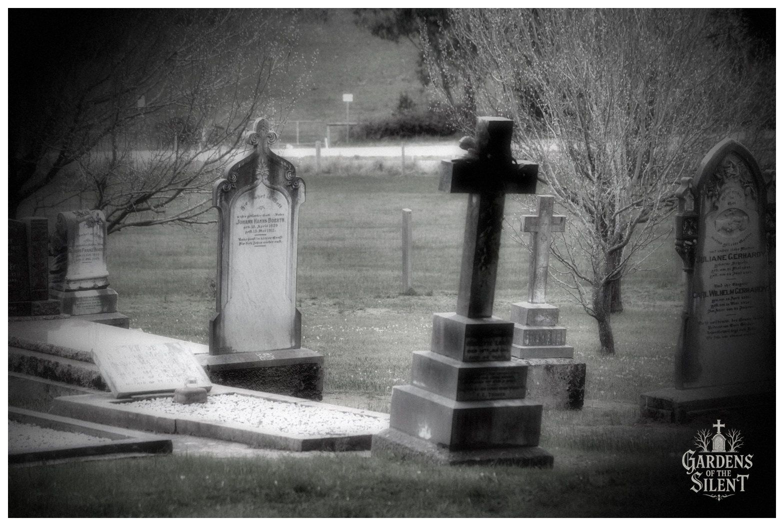 Atmospheric black and white photograph depicting a collection of historic headstones set amongst young deciduous trees with fine bare branches. In the foreground stands a tall, slightly tilted stone cross on a stepped pedestal. To the left is an ornate arched headstone for Johann Hanns Boerth, and to the right, a darker arched monument for Juliane Gerhardy. A soft, hazy light blankets the open grass and distant background. The Gardens of the Silent watermark appears in the bottom right corner.