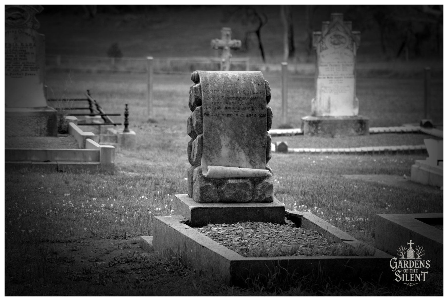 Black and white photograph of an intricately sculpted headstone designed as an open stone scroll draped over rough hewn ashlar rockwork. The inscription commemorates Ernestine W. Seiler, née Zimmermann, who died in 1912. The monument sits on a dark base with a stone kerb filled with gravel in the foreground. In the softly blurred background, other historic markers and distant trees create a tranquil cemetery setting. The watermark Gardens of the Silent is present in the lower right corner.