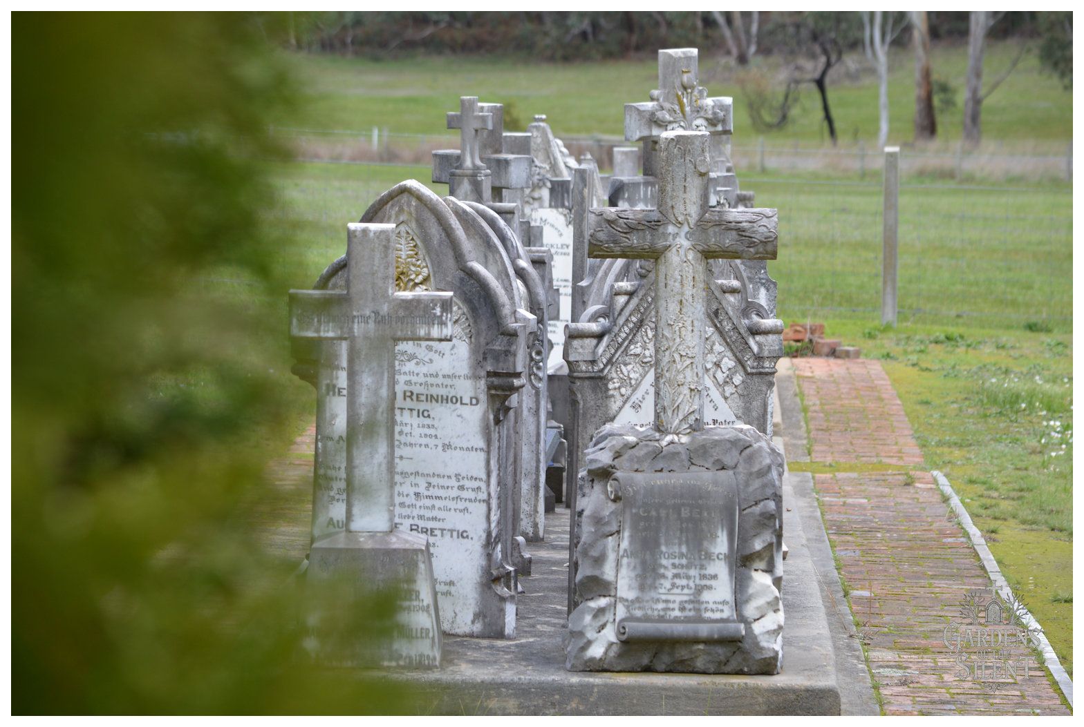 Colour photograph showing a neat row of historic stone headstones and crosses standing along a concrete base. The foreground features a weathered grey cross intricately carved with climbing ivy, resting on a rustic boulder base with an unrolled scroll inscription for Carl and Anna Rosina Beck. Behind it, several matching arched and cross headstones recede in perspective. A blurred green bush frames the left edge, while a red brick walking path borders the graves on the right, leading towards open green paddocks. The Gardens of the Silent watermark is visible in the lower right corner.