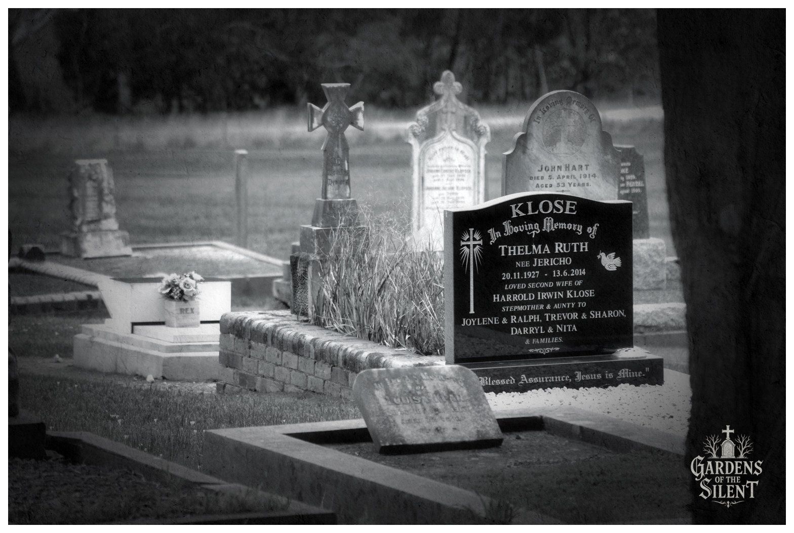 Black and white photograph of a cemetery plot displaying a blend of modern and historic headstones. In the midground, a polished black granite headstone stands within raised brick kerbing overgrown with tall ornamental grass, inscribed for Thelma Ruth Klose, who died in 2014. Older weathered white marble markers surround the plot, including a Celtic cross, an arched headstone for John Hart from 1914, and a low stone base with a flower vase inscribed Rex. A dark tree trunk frames the right edge of the frame against a soft focus background. The Gardens of the Silent logo is visible in the lower right corner.
