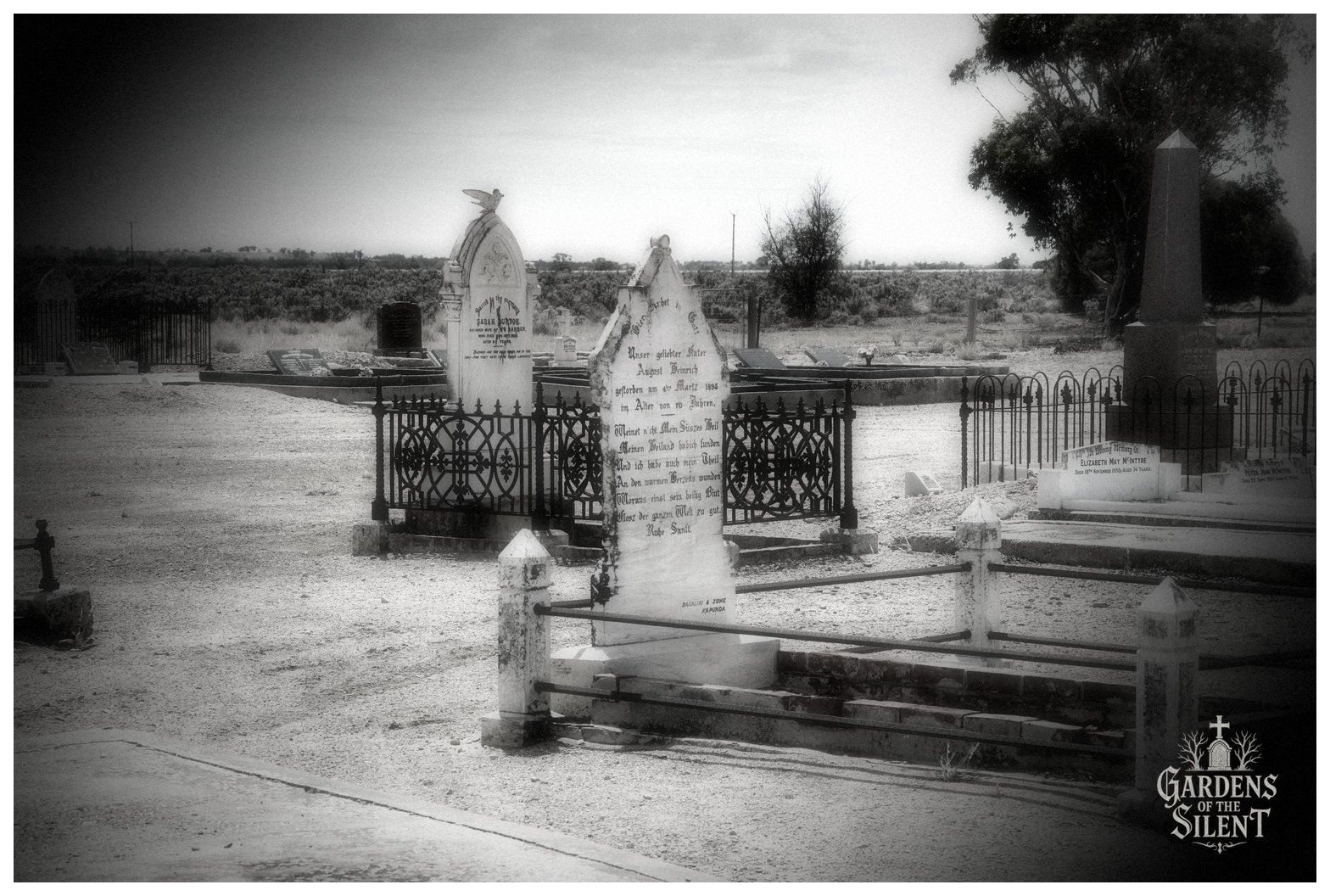 A moody black and white photograph framed with a dark vignette. In the foreground stands a pointed marble headstone with German inscriptions, surrounded by intricate decorative wrought iron railings on low concrete footings. In the background, additional monuments, including a carved dove atop a marker and a tall dark obelisk, stand across the dusty ground against a low line of native scrub.