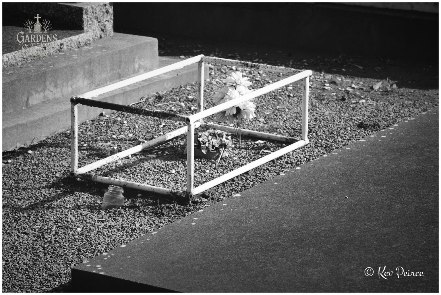 A high contrast black and white composition focusing on a small, low white metal pipe frame surrounding a gravel plot. Inside the frame, a simple arrangement of faux flowers rests beside a small glass jar on the gravel. Sunlight illuminates the white piping against the smooth dark stone kerbing in the foreground.