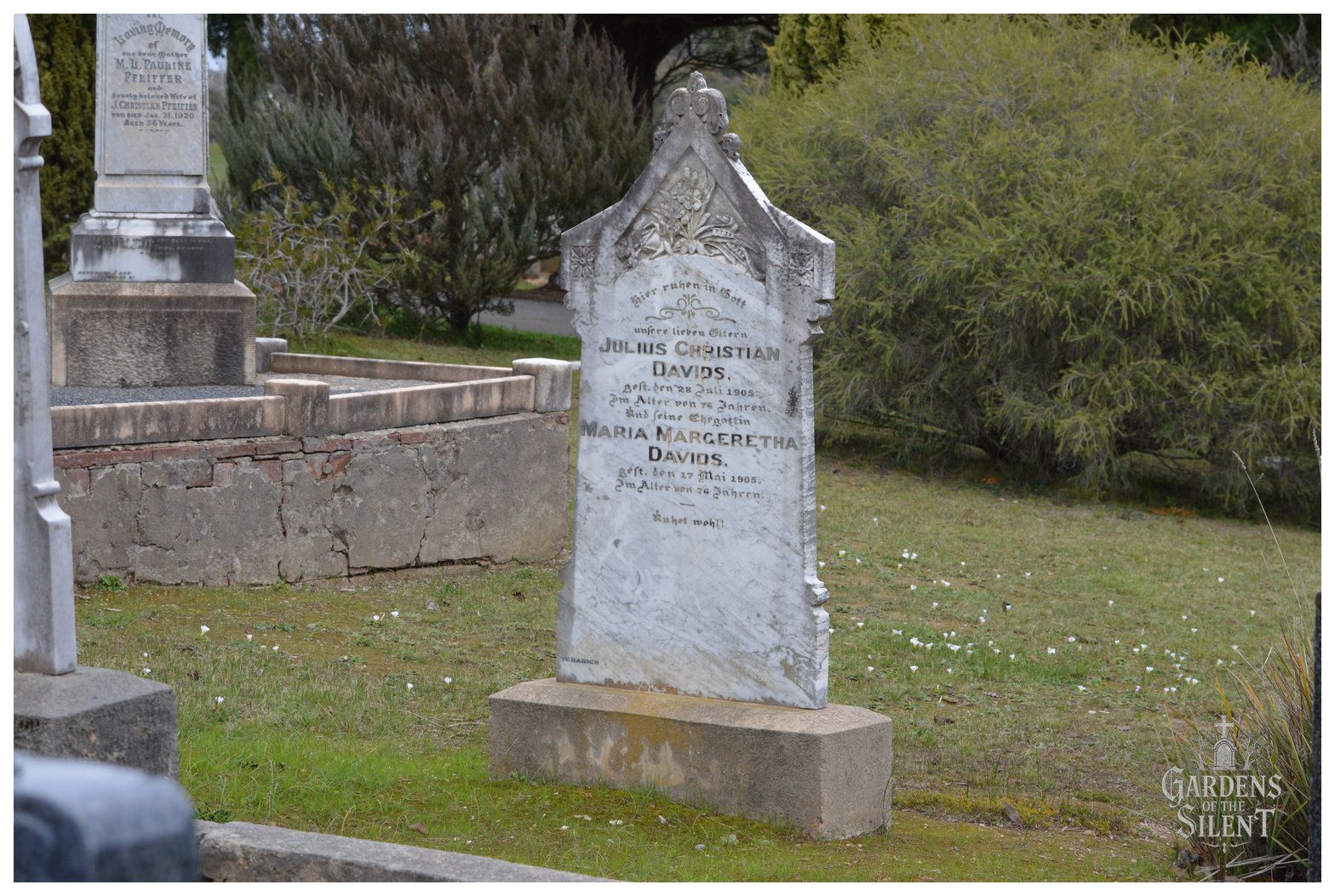 Colour photograph featuring an upright marble headstone with an ornate gabled top and carved floral relief. The inscription in German honours Julius Christian Davids and his wife Maria Margeretha Davids, both of whom passed away in 1905 at the age of 76. The weathered stone rests on a sand coloured plinth set in an open lawn scattered with tiny white flowers. To the left is an elevated, stone walled grave plot, with a large native bush filling the background. The Gardens of the Silent watermark rests in the lower right corner.